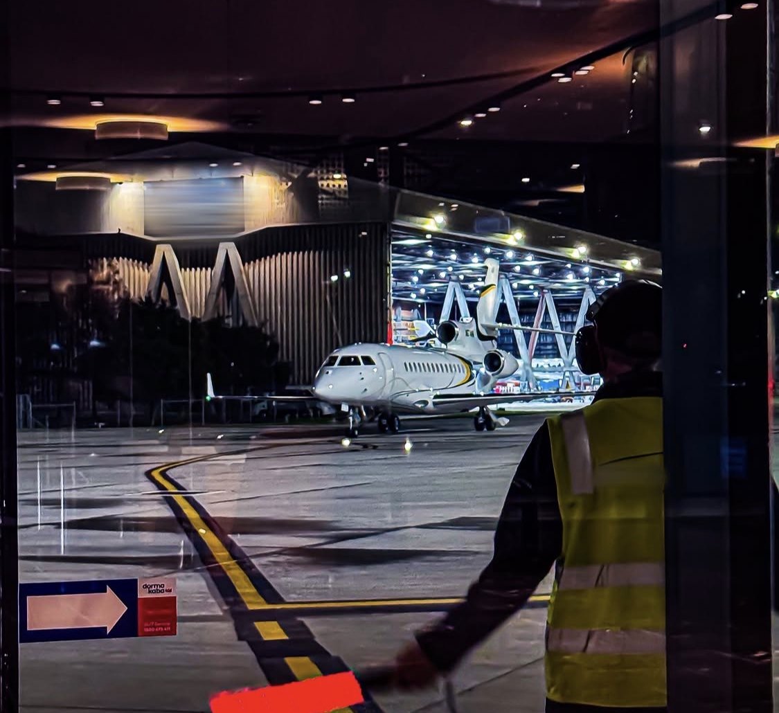 An aircraft on a floodlit apron at night, seen through the glass of a terminal, with ground crew alongside.