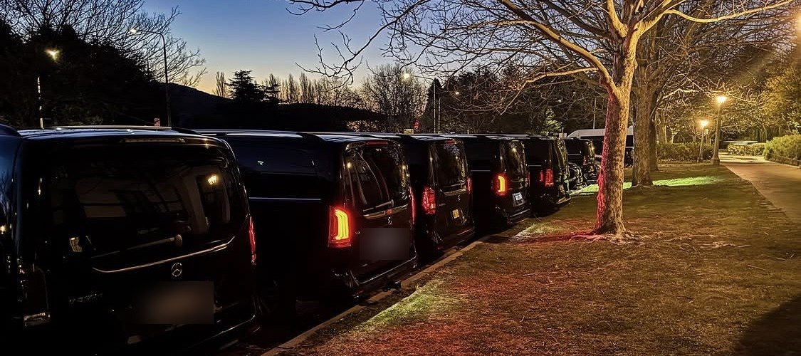 A line of black protection vehicles staged under trees at dusk, lights on, waiting to move.