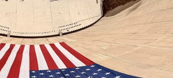 The Hoover Dam from above, a large United States flag laid across the apron below it.