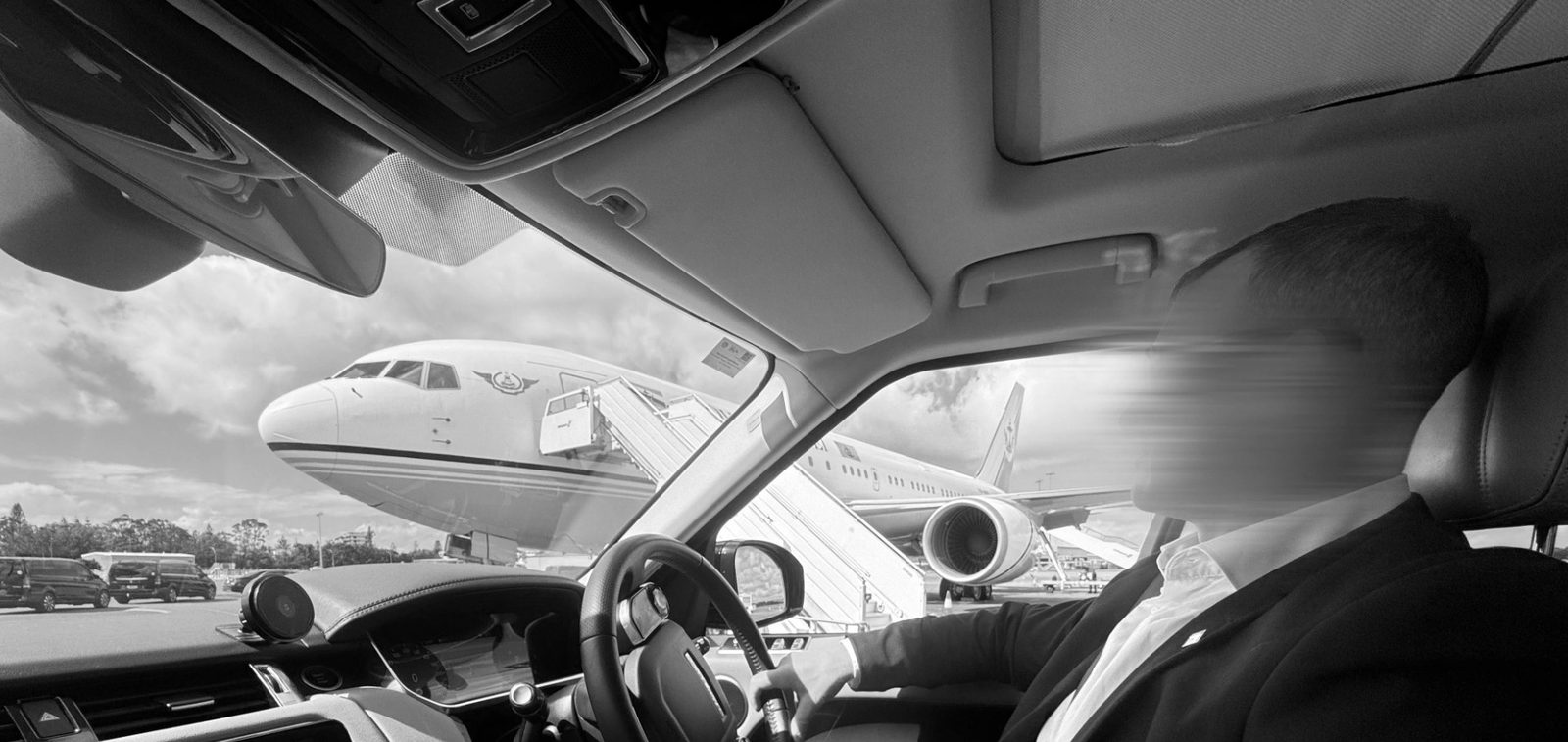 A security driver at the wheel of a vehicle on an airport apron, aircraft waiting on the tarmac beyond the windscreen.